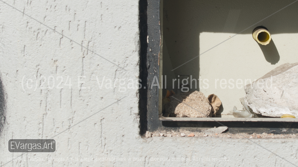 a square hole on a white street wall, plastic pipe, small rocks inside, shadows, sunlight, outdoors, day, daylight, street, urban